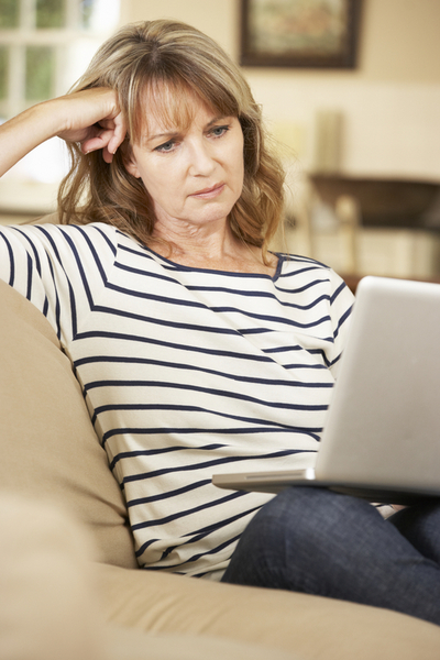 Puzzled Mature Woman Sitting On Sofa At Home Using Laptop